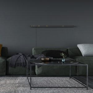 Wohnzimmer mit dunkler Einrichtung und moderner Wandgestaltung. An der Wand ist eine längliche Leuchtschrift-Uhr montiert, die gelb beleuchtete Wörter anzeigt. Darunter steht ein dunkelgrünes Sofa mit grauem und schwarzem Kissen sowie einer dunklen Decke. Davor befindet sich ein schwarzer Couchtisch mit dünnem Metallrahmen, auf dem eine kleine Vase mit Zweig und eine Schale stehen. Der Boden ist mit einem grauen Teppich bedeckt. Die Szene wirkt minimalistisch und harmonisch abgestimmt.